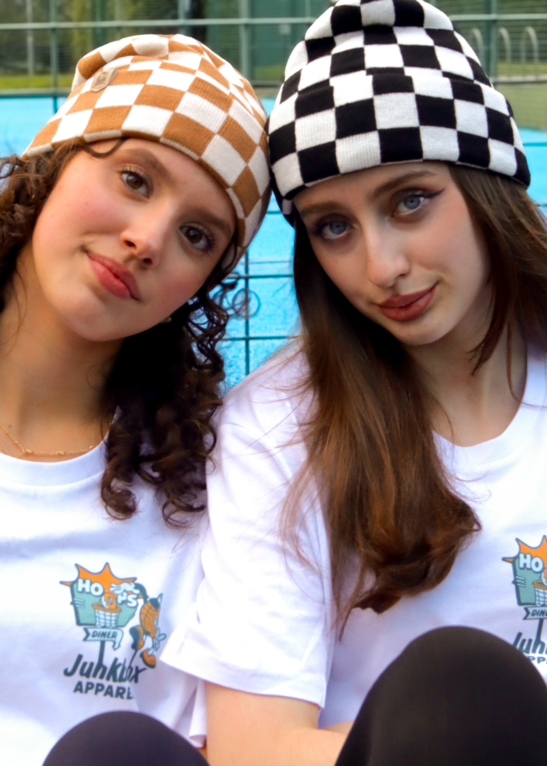Two young women sitting together in front of a tennis court, wearing matching white t-shirts with a 'Junkbox Apparel' graphic and checkered beanie hats.