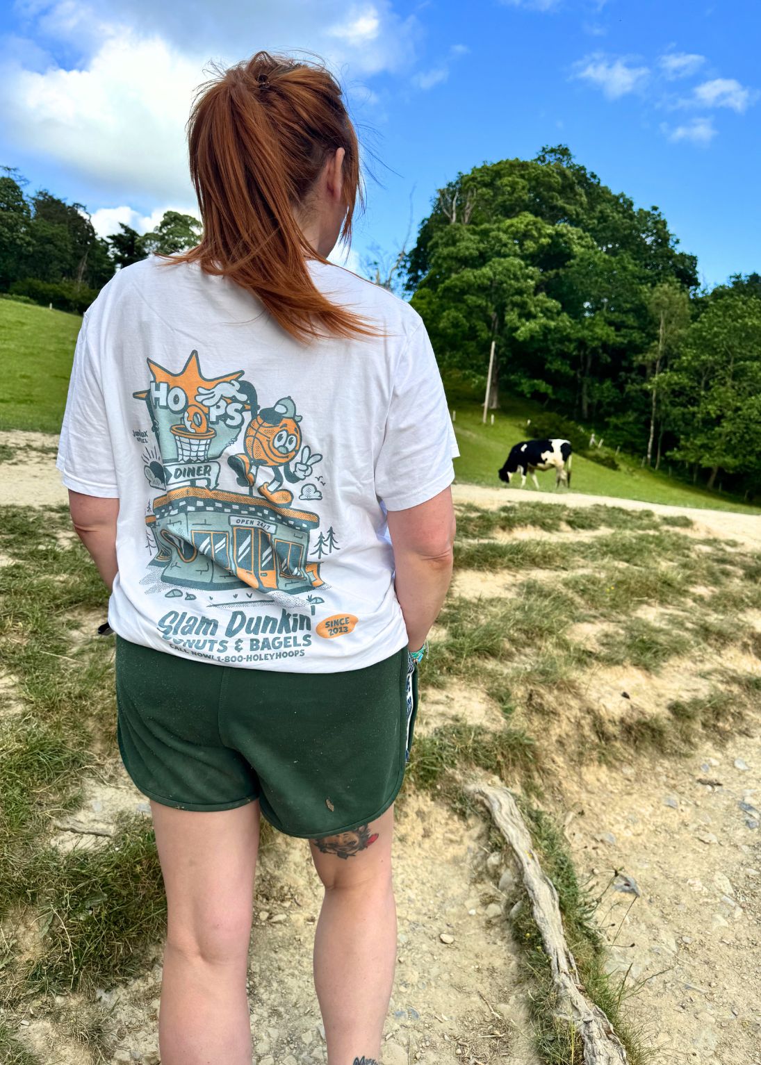 A woman viewed from behind wearing a white graphic t-shirt with a colorful cartoon basketball design and green athletic shorts, standing outdoors on a path with trees, grass, and a cow in the background.