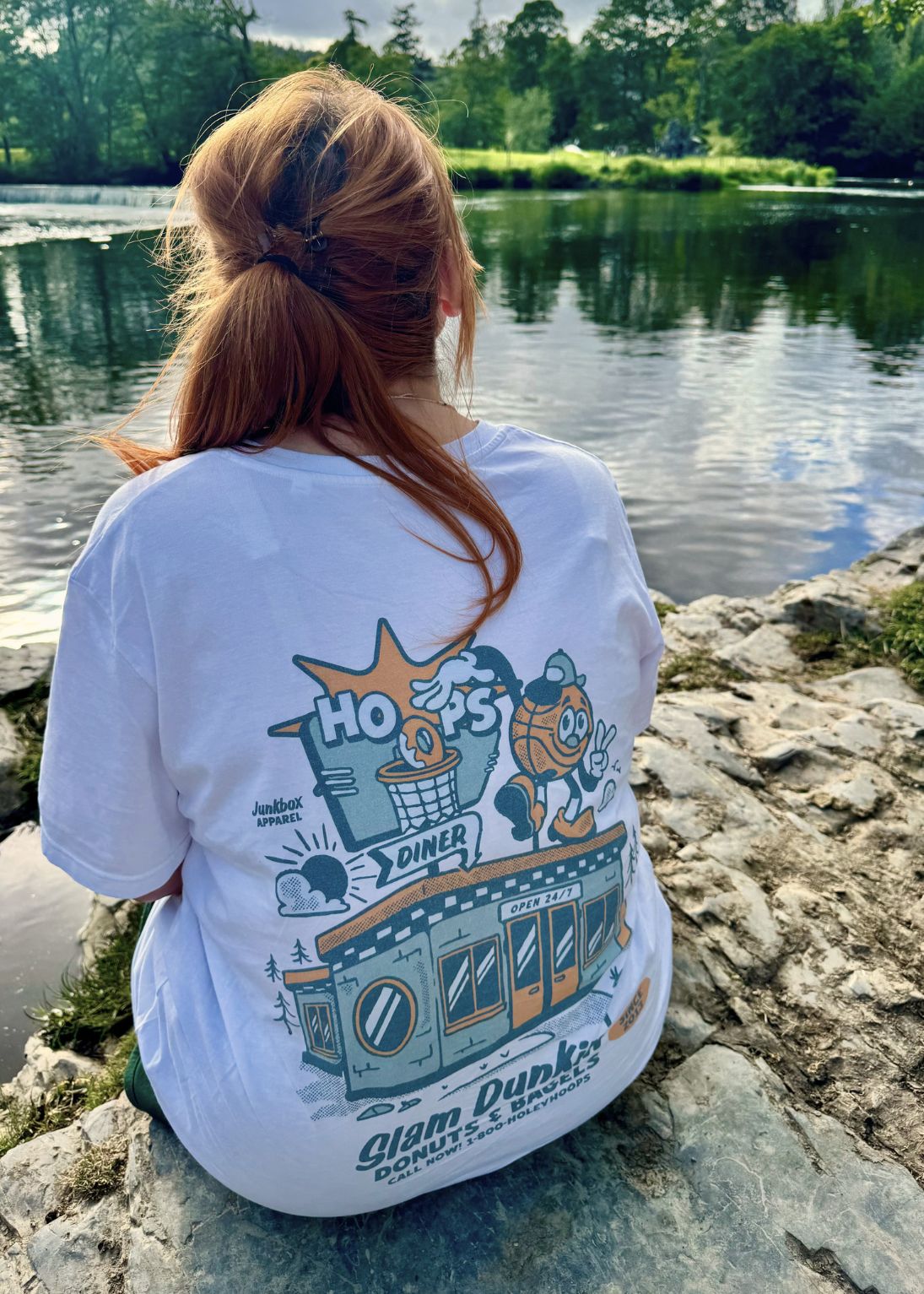 A person sitting on rocks by a lake, viewed from behind, wearing a white graphic T-shirt with a large illustrated back print reading 'Hoops Diner' and 'Slam Dunkin'.