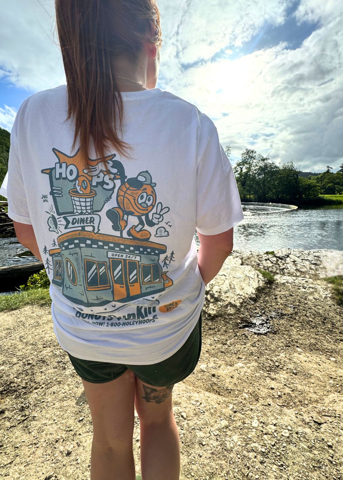 A person standing outdoors by a body of water, viewed from behind, wearing a white graphic T-shirt with a large multicolor basketball-themed print and dark green shorts.