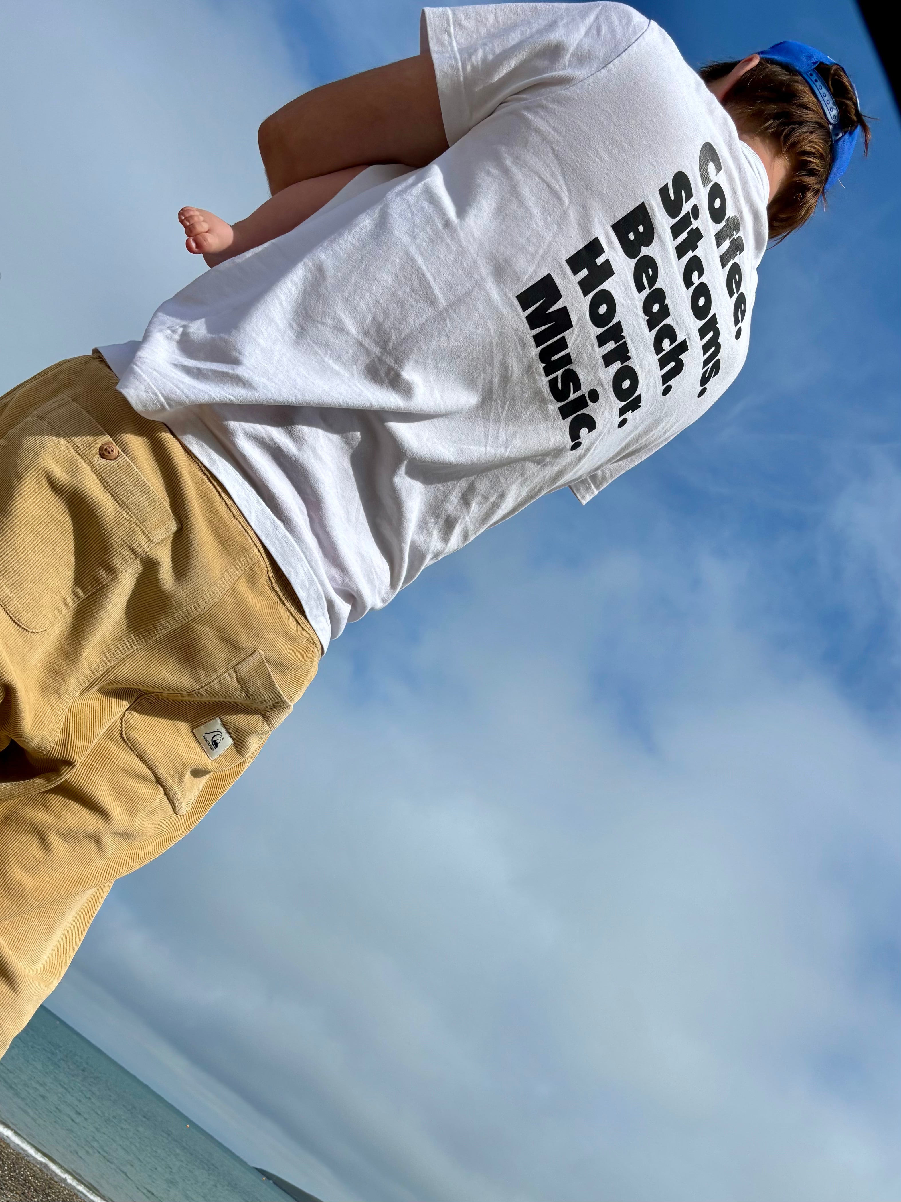 A person outdoors wearing a white graphic T-shirt with vertical black text reading 'Coffee. Sitcoms. Beach. Horror. Music' and tan pants, photographed from a low angle against a blue sky.