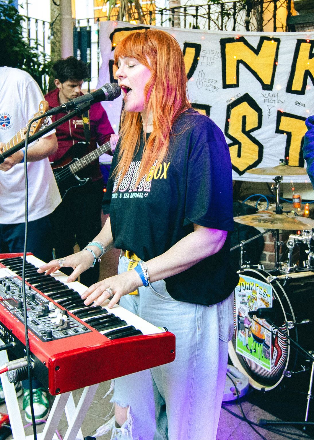 Woman with red hair playing keyboard and singing at indoor music event, wearing black graphic t-shirt and ripped jeans.