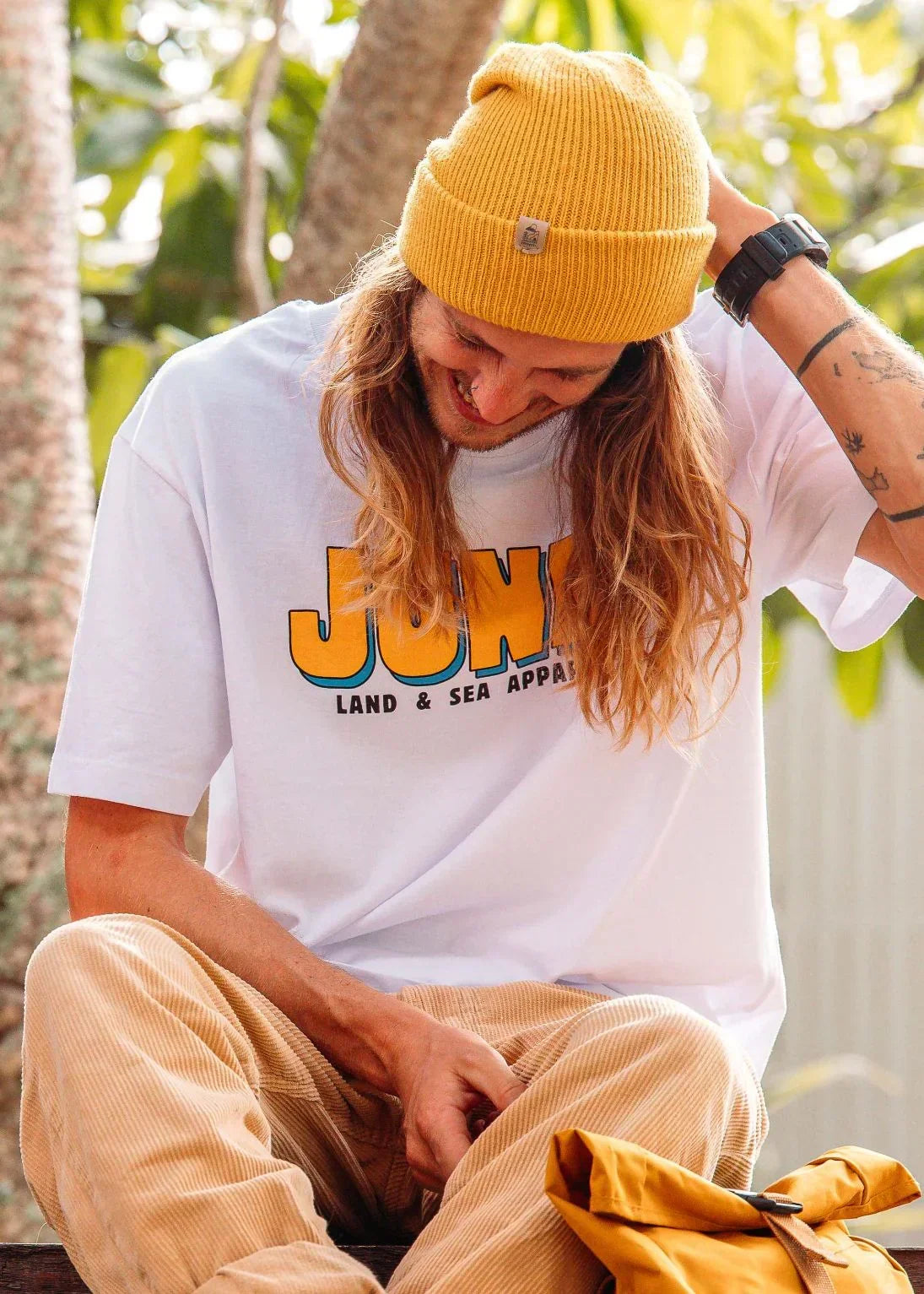 Smiling person in a yellow beanie and white t-shirt with 'Land & Sea Apparel' text, outdoors