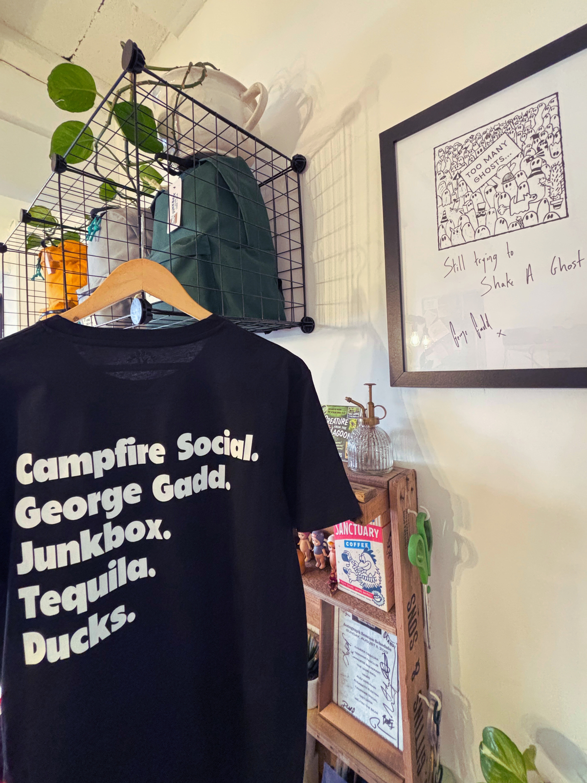 A black short-sleeve graphic T-shirt hanging on a wooden hanger in a shop interior. The shirt has white text reading 'Campfire Social.