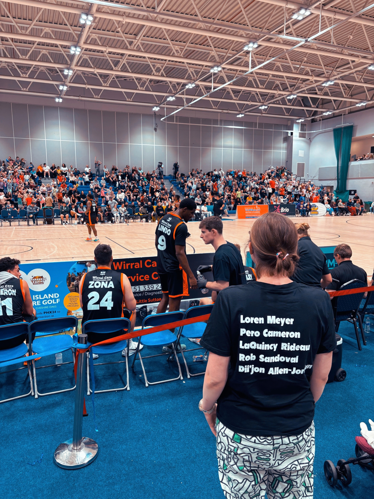 A person wearing a black graphic t-shirt with the names of basketball legends printed on the back stands on a blue carpeted indoor sports court, facing a basketball game in progress. The background shows a crowded indoor arena with spectators in the stands.