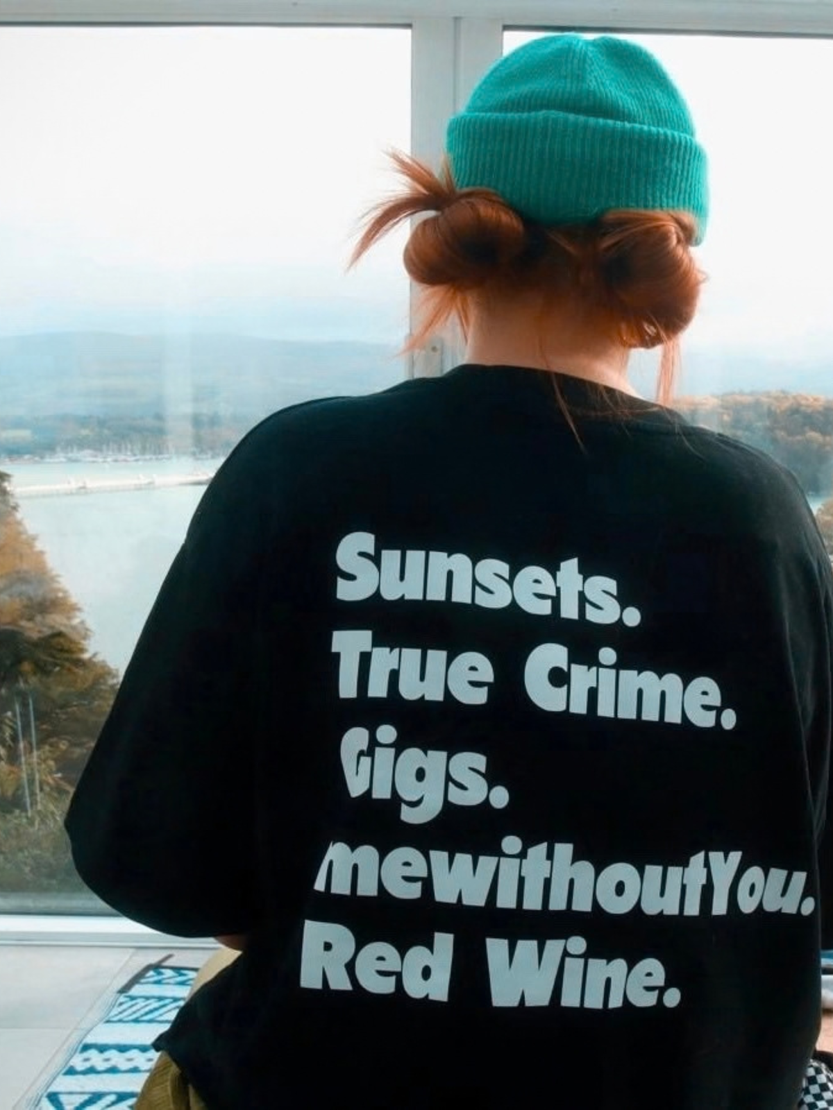 A person viewed from behind wearing a black oversized t-shirt with white text that reads 'Sunsets. True Crime.