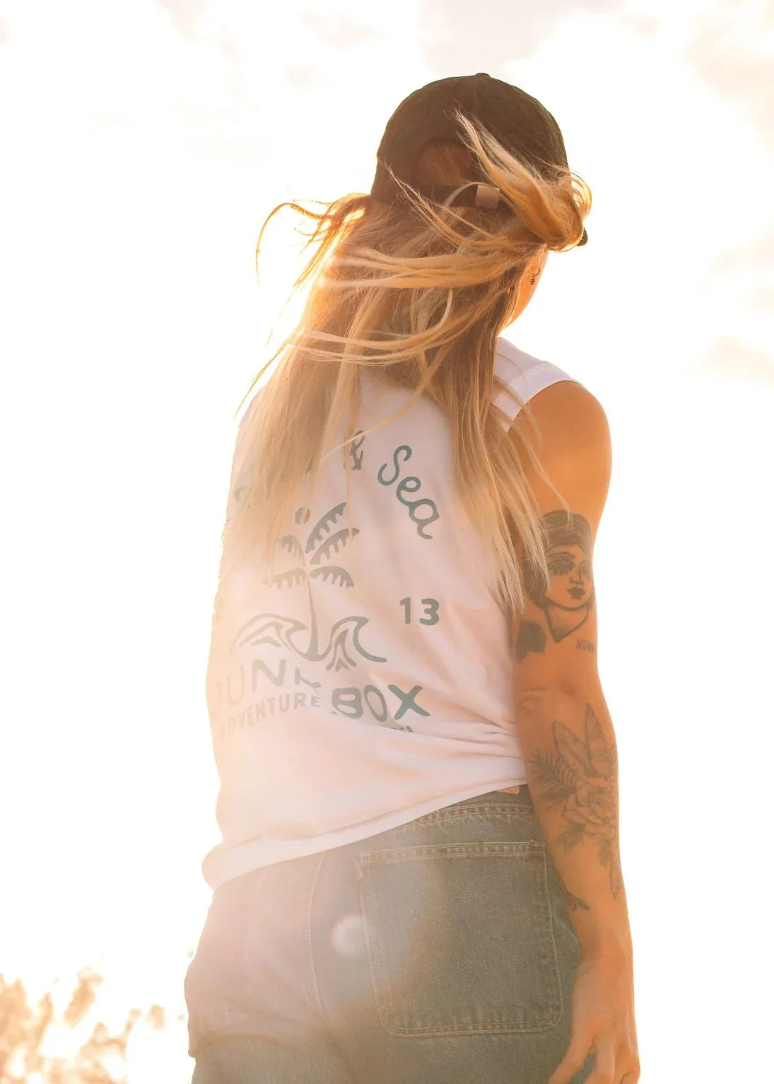 Woman in eco-friendly tank top with 'Junk Box Adventure' print, facing sunset, tattoos visible