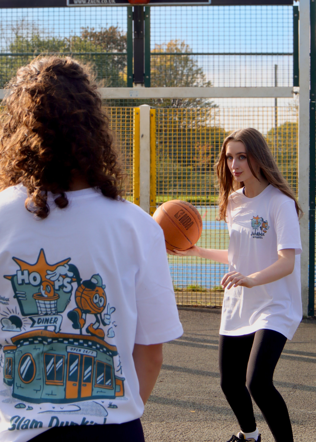 Two people on an outdoor basketball court wearing matching white graphic t-shirts. The person on the left shows the back of the shirt with a colorful cartoon graphic, and the person on the right holds a basketball while wearing the front of the matching shirt.