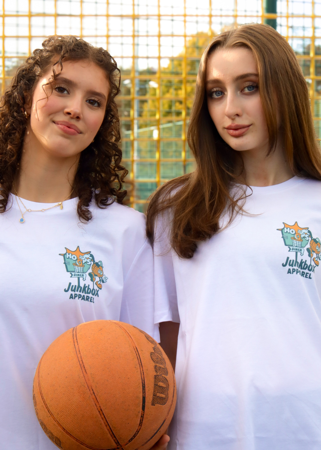 Two young women standing side-by-side in front of a yellow basketball court net, wearing matching white Junkbox Apparel t-shirts and holding an orange basketball.