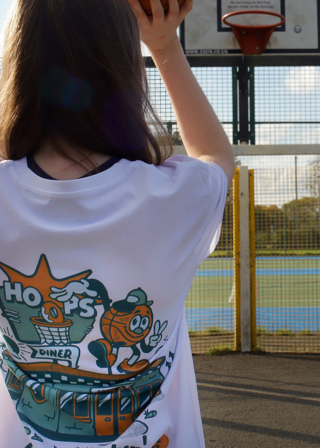 A person viewed from behind wearing a white graphic t-shirt with a colorful cartoon design on the back, standing on an outdoor basketball court with a hoop in the background.