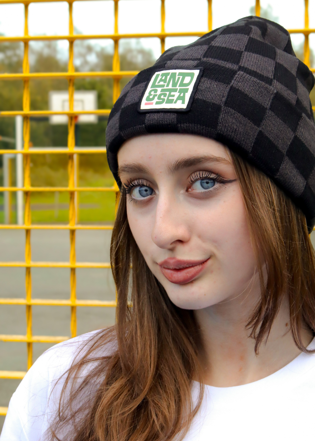 A close-up portrait of a woman wearing a black and dark gray checkered beanie with a green and white 'LAND & SEA' patch on the front, set against a bright yellow metal fence.