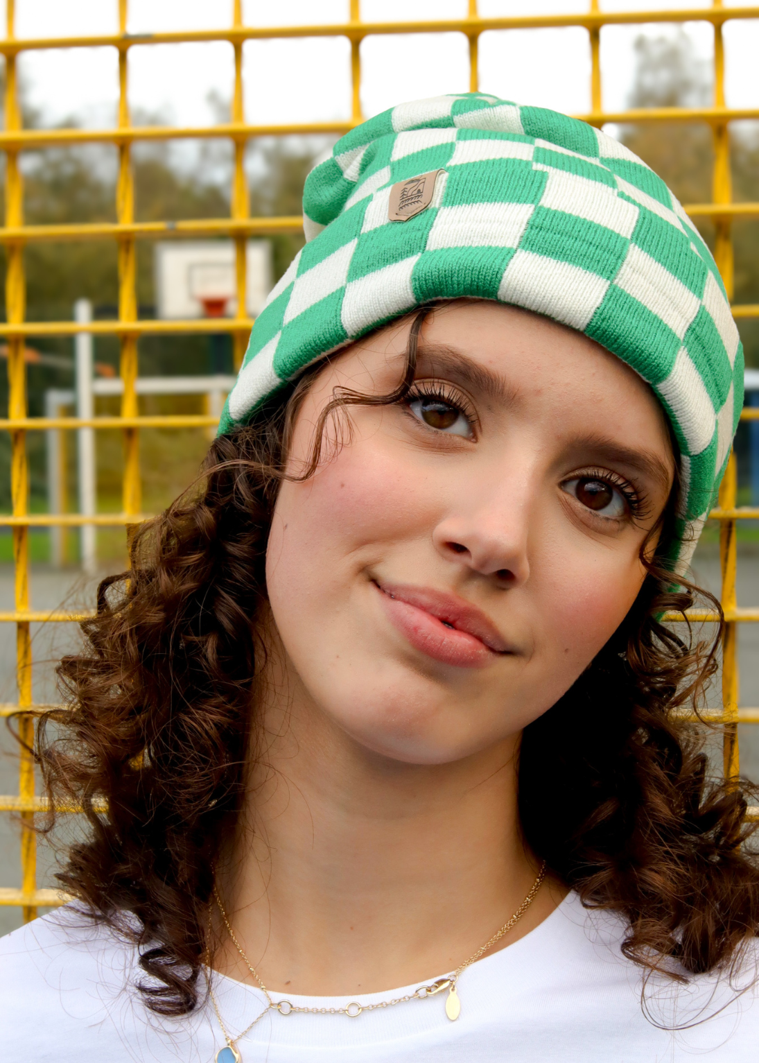 A close-up portrait of a woman wearing a green and white checkered knit beanie hat, standing in front of a yellow metal fence.