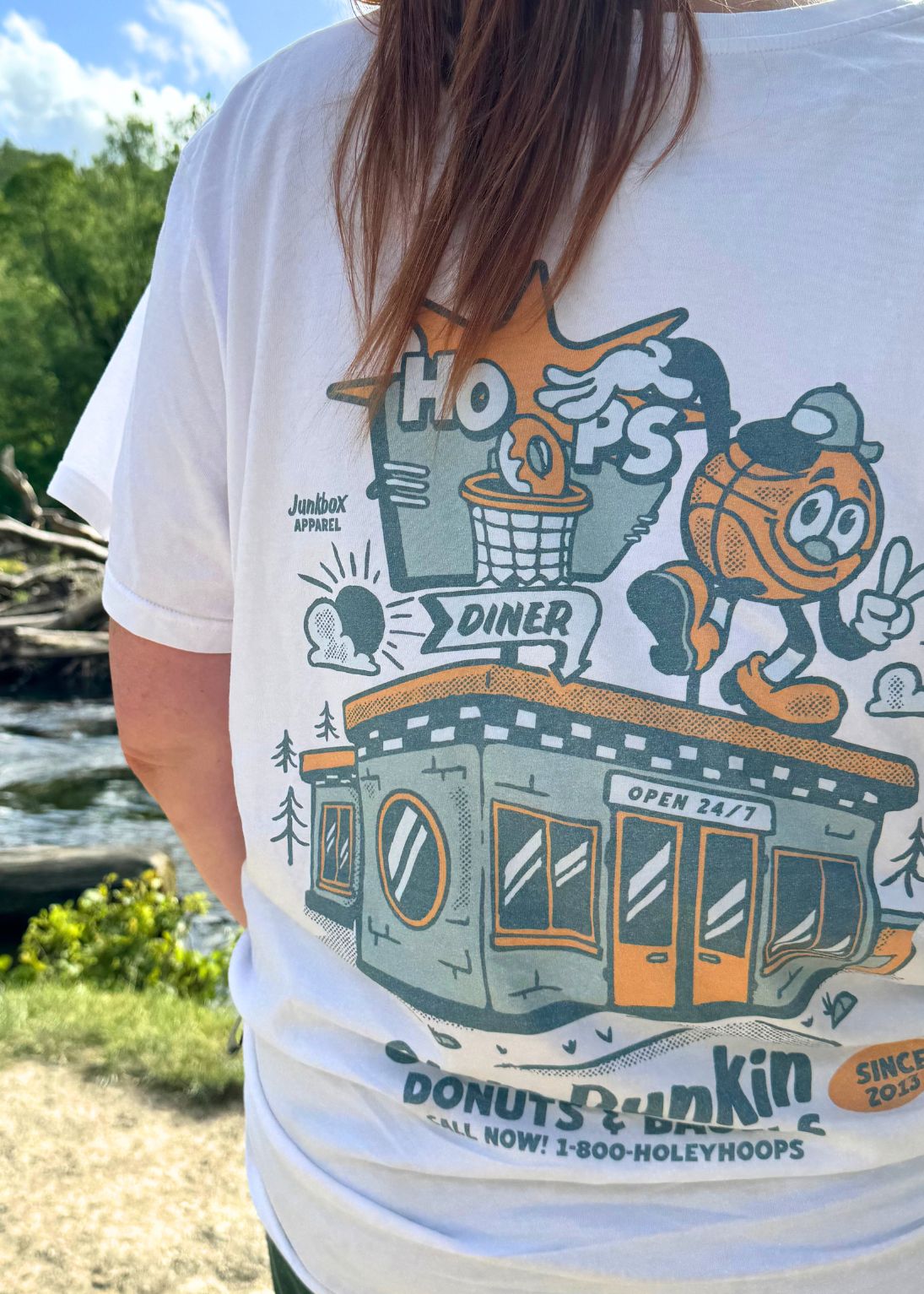 A person outdoors wearing a white graphic T-shirt with a large retro donut-themed print featuring a diner, basketball mascot, and text including 'Hoops' and 'Donuts Dunkin''.