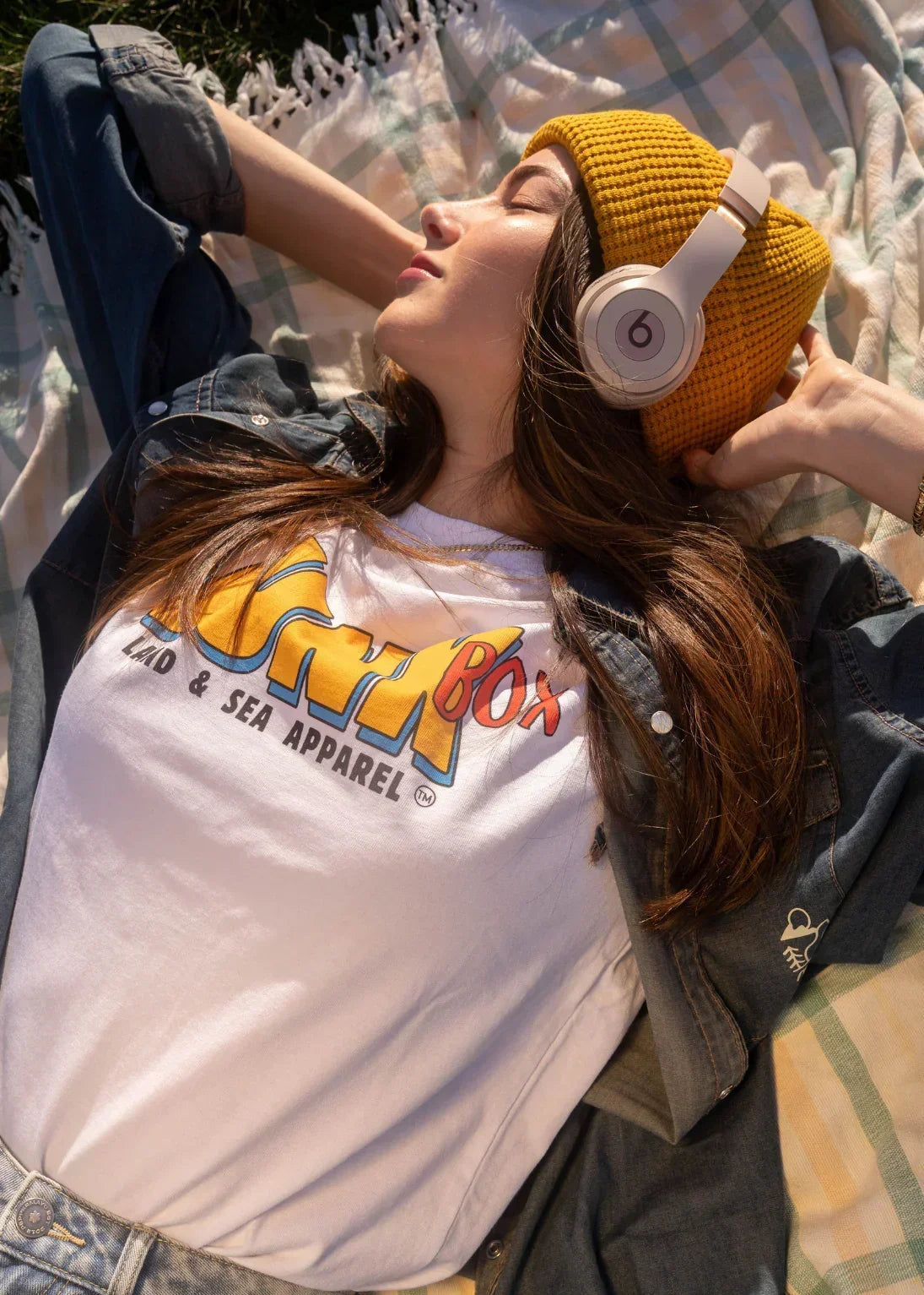 Woman relaxing outdoors wearing yellow beanie, headphones, and graphic eco friendly T-shirt