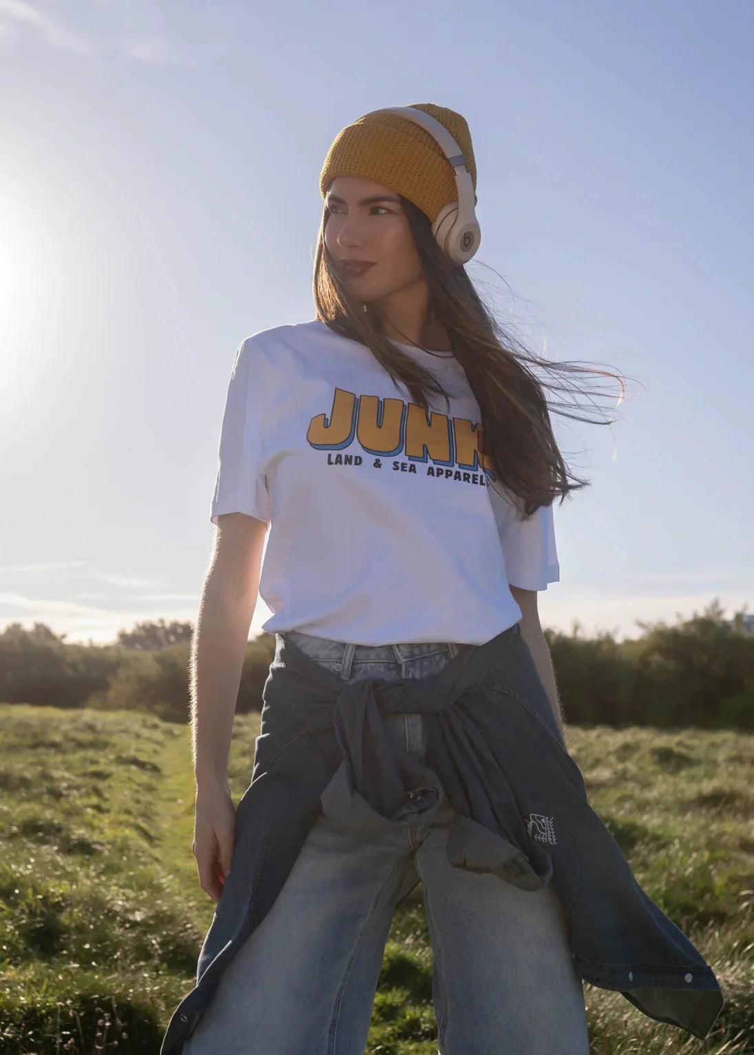 Woman in yellow beanie and headphones wearing white graphic t-shirt outdoors in sunlight