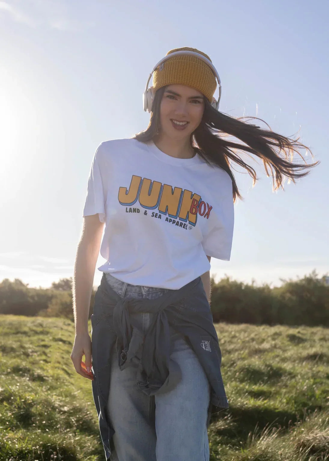 Woman outdoors wearing eco-friendly graphic t-shirt, yellow beanie, headphones in grassy field