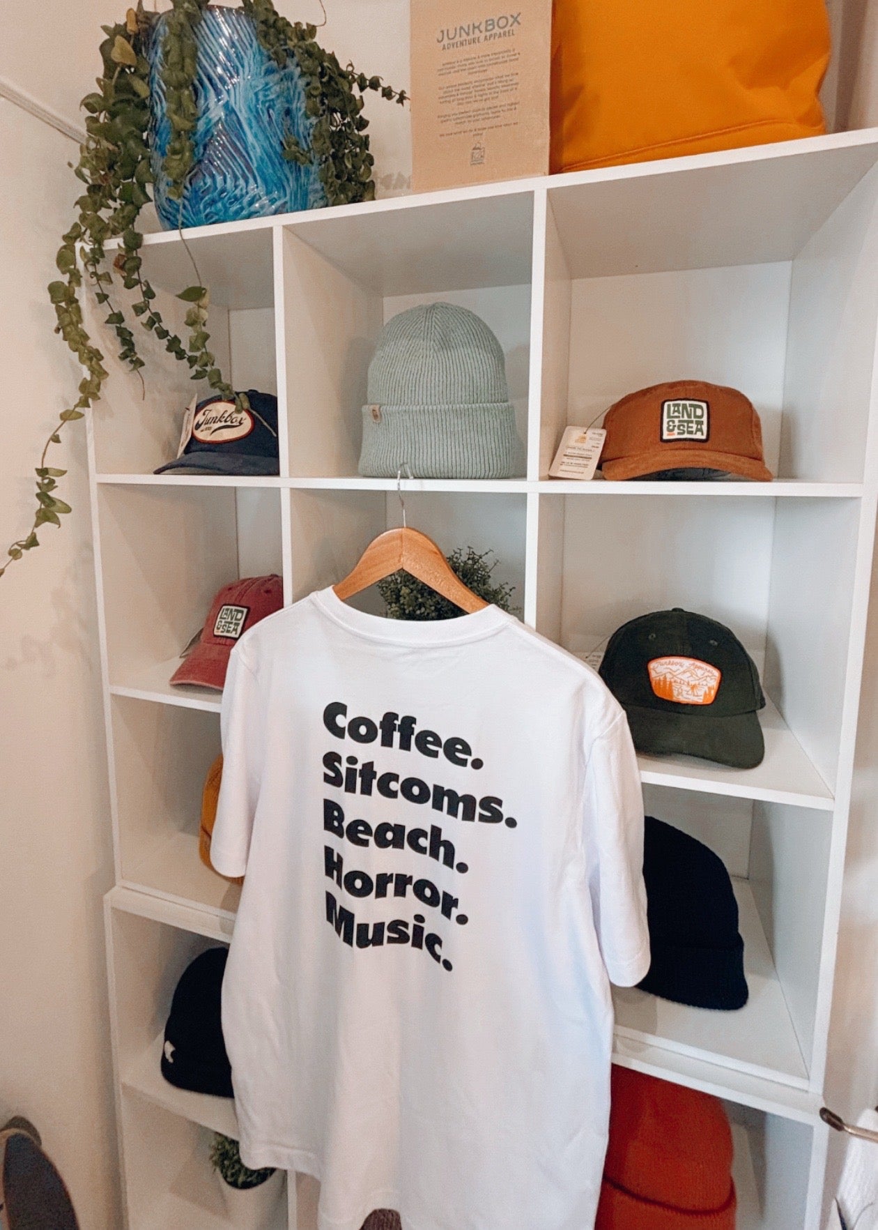 White t-shirt with text on a white shelf with hats and a plant.