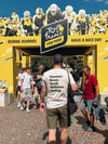 People walking past a Tour de France fan park entrance with fans and cyclists in the background.