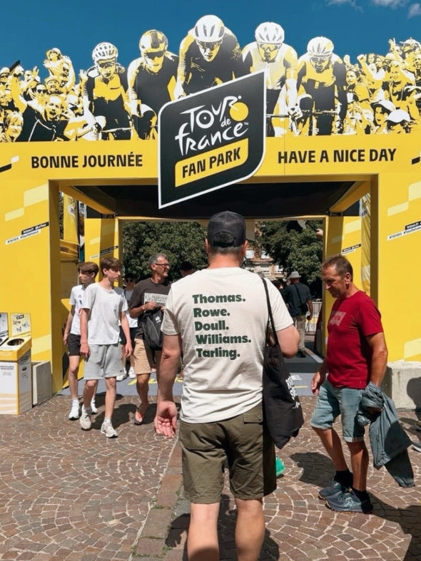 People walking past a Tour de France fan park entrance with fans and cyclists in the background.