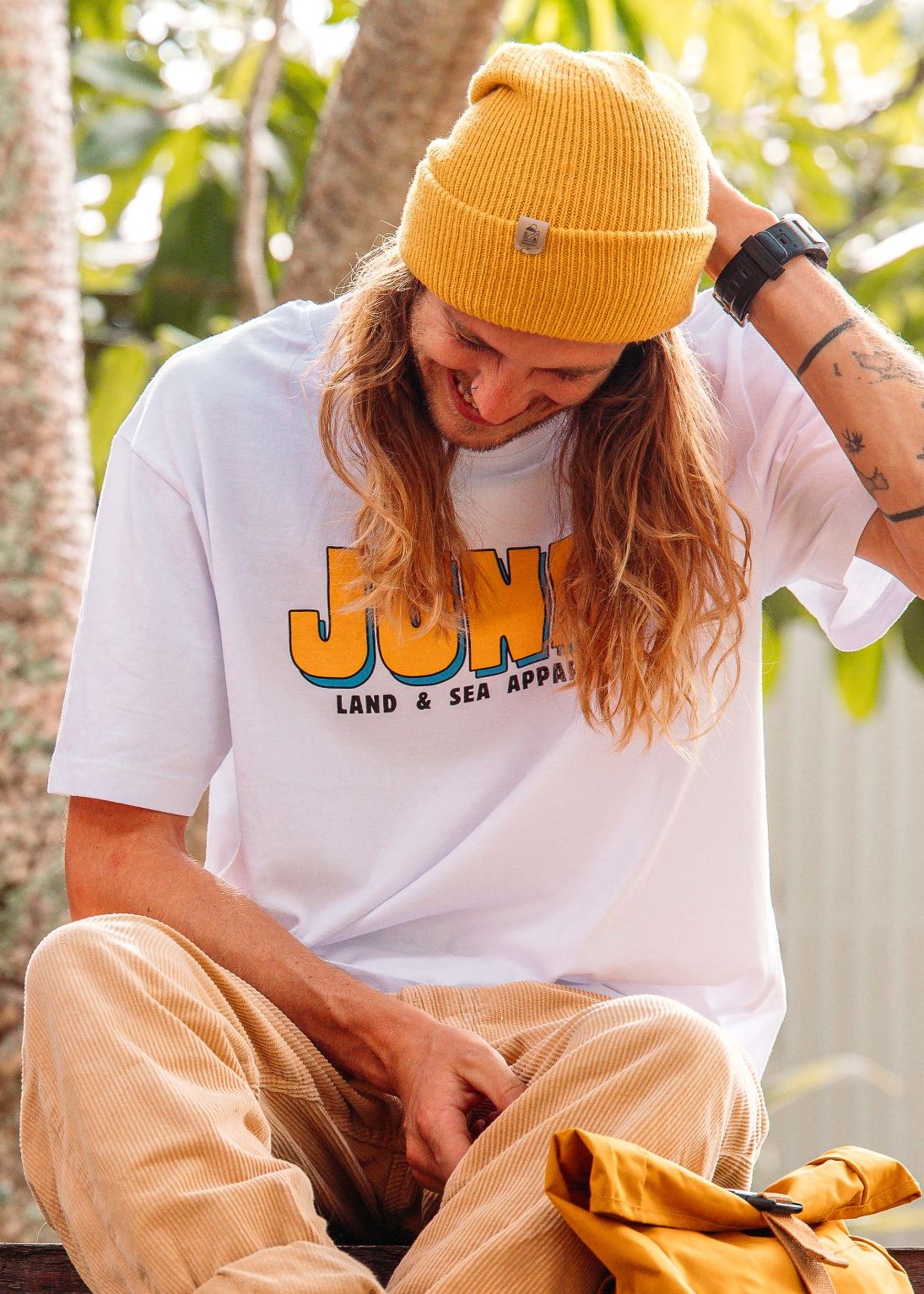 Person wearing a white t-shirt with 'JUNK' branding, yellow beanie, and beige pants sitting outdoors.
