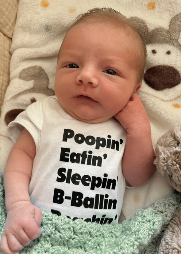 Newborn baby wearing a onesie with humorous text, lying on a soft surface with a plush toy.