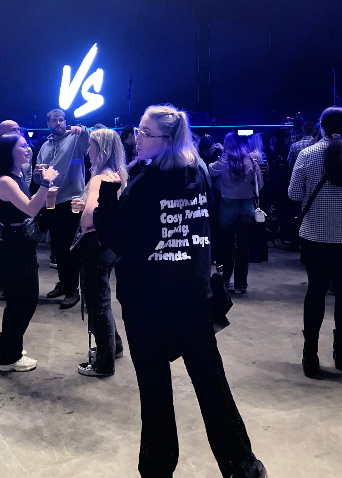 Person wearing a black hoodie with text at a social event with 'VS' neon sign.