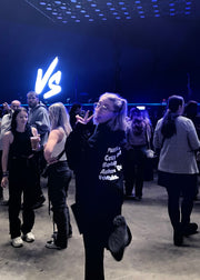 Person wearing a junkbox hoodie posing in front of a 'VS' neon sign with people around
