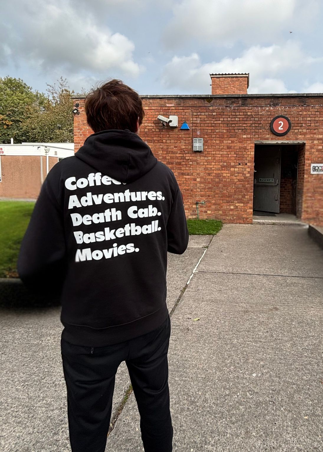Person wearing a black hoodie with text on the back, standing in front of a brick building.