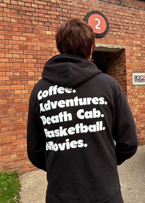 Person wearing a black hoodie with text on the back in front of a brick wall.