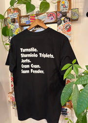Black t-shirt with white text on a hanger against a wall with various stickers and plants.