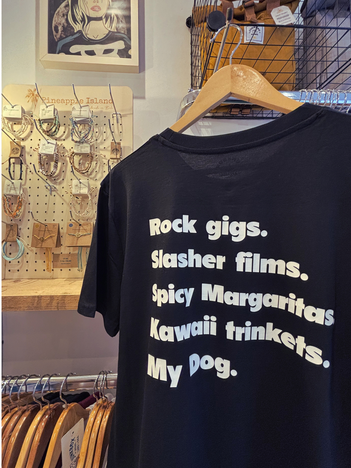 A black graphic t-shirt hanging on a wooden hanger in a retail store. The shirt features white text listing phrases like 'Rock gigs', 'Slasher films', 'Spicy Margaritas', 'Kawaii trinkets', and 'My dog'.