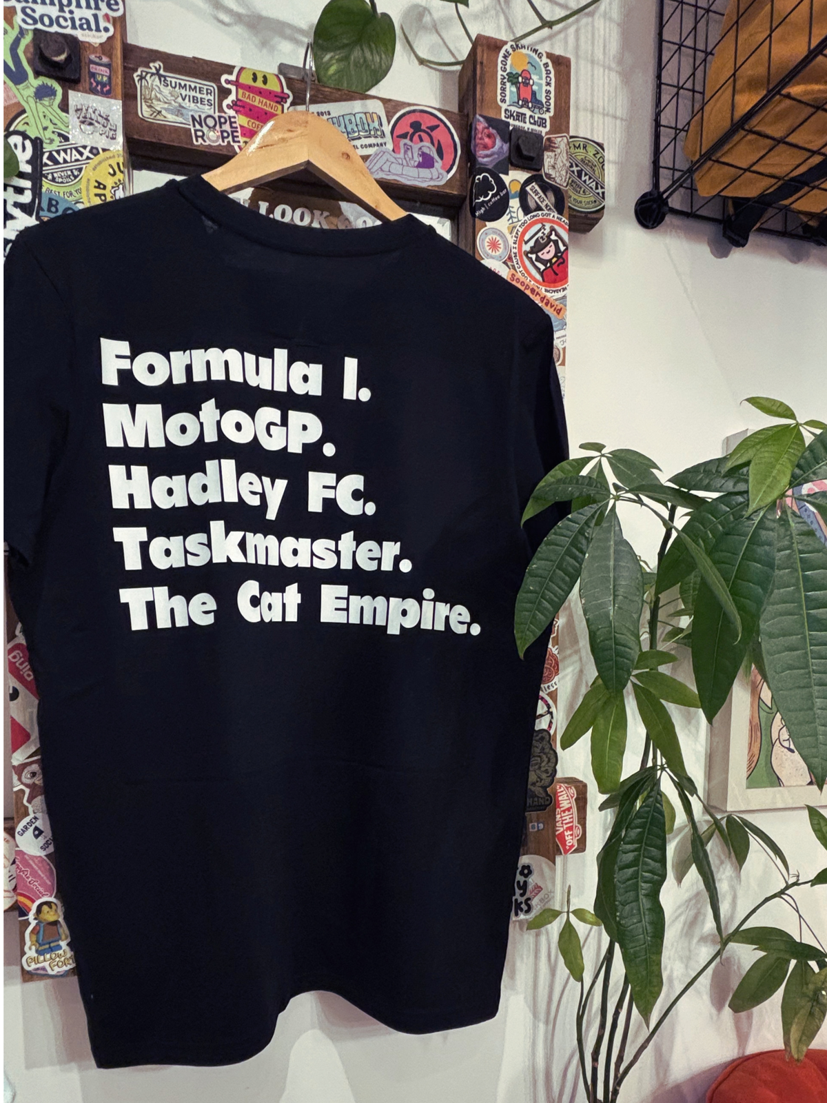 A black t-shirt hanging on a wooden hanger against a white wall decorated with stickers and a green plant. The shirt features white text listing: 'Formula 1, MotoGP, Hadley FC, Taskmaster, The Cat Empire.