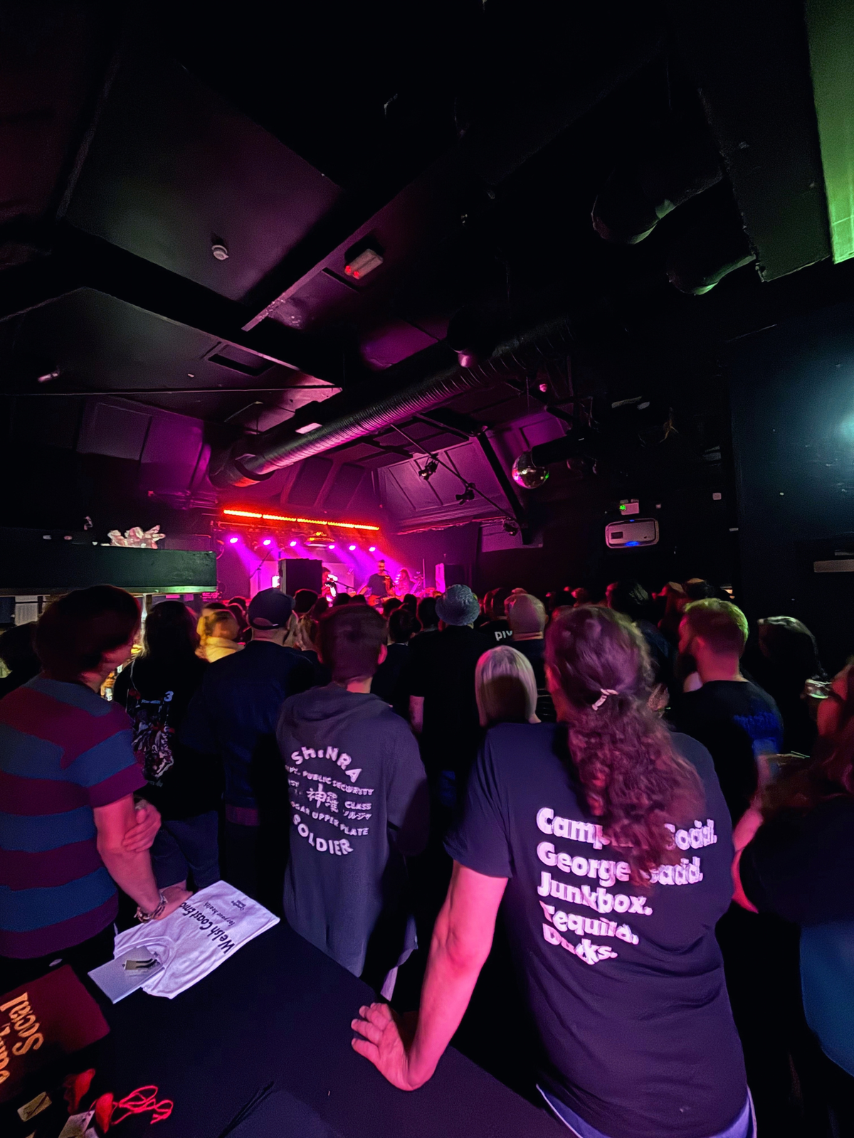 A crowd of people viewed from behind in a dimly lit venue with purple stage lighting, wearing dark t-shirts with white printed text. One shirt reads 'Campfire Social George Gadd Junkbox Tequilla Ducks'