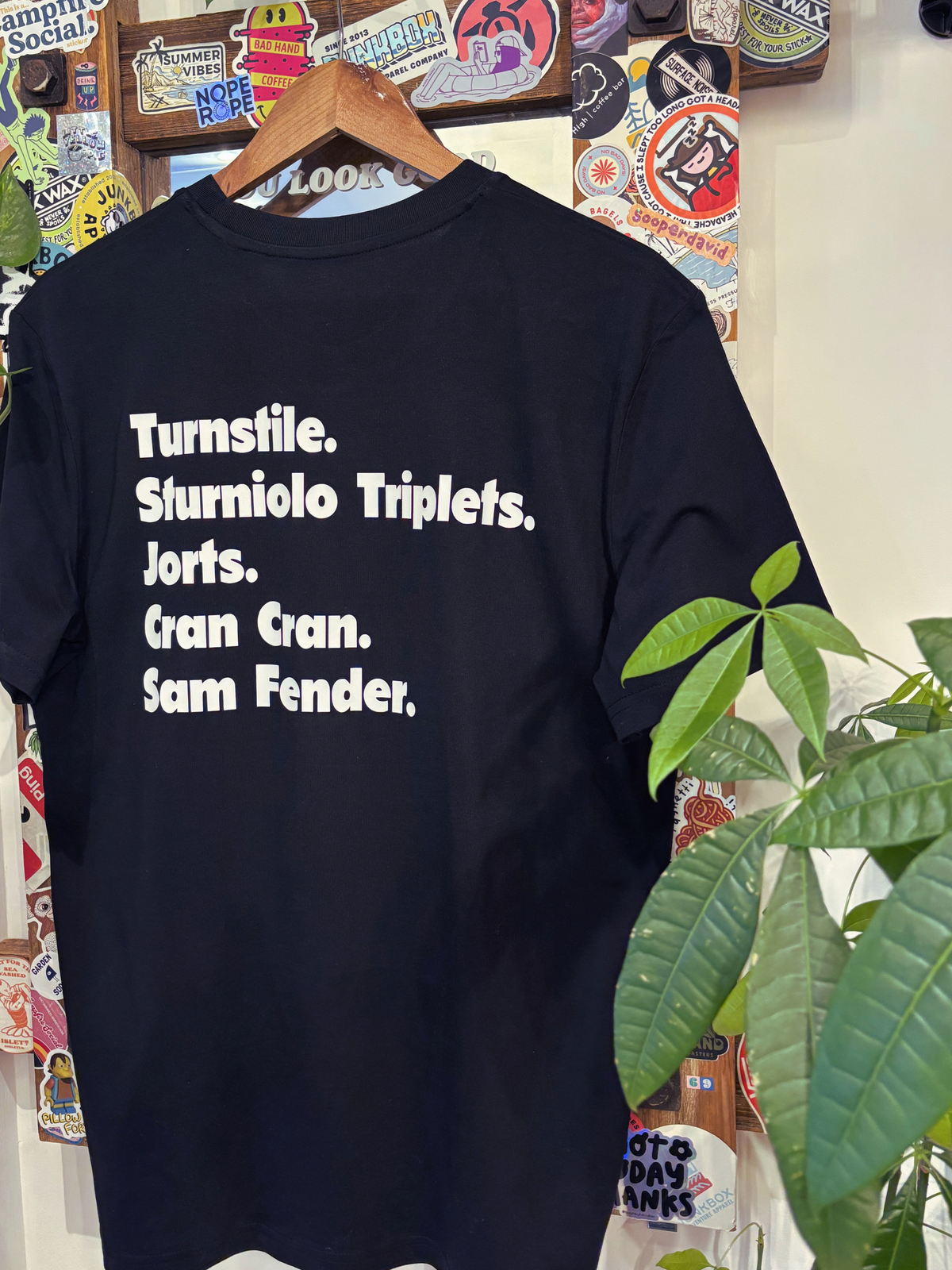 A black short-sleeve graphic t-shirt hanging on a wooden hanger against a wall covered in stickers, with a green plant in the foreground. The shirt features white text listing names and terms: 'Turnstile, Sturniolo Triplets, Jorts, Gran Gran, Sam Fender.