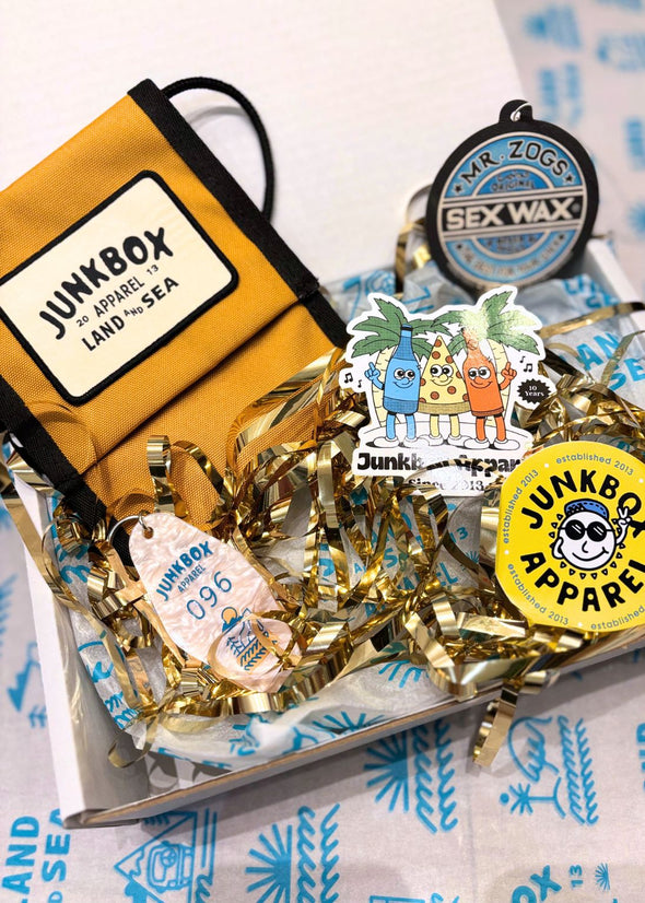 Collection of Junkbox branded items including a yellow bag, stickers, and a sex wax air freshener on a patterned surface.