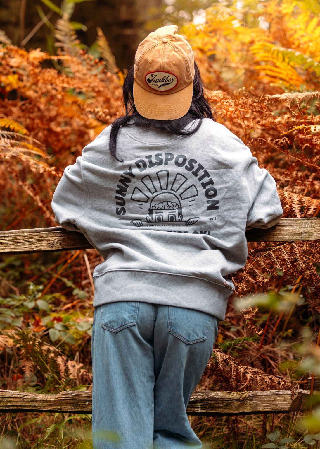 Person wearing a cap and sweatshirt with text, leaning on a log in a forest setting
