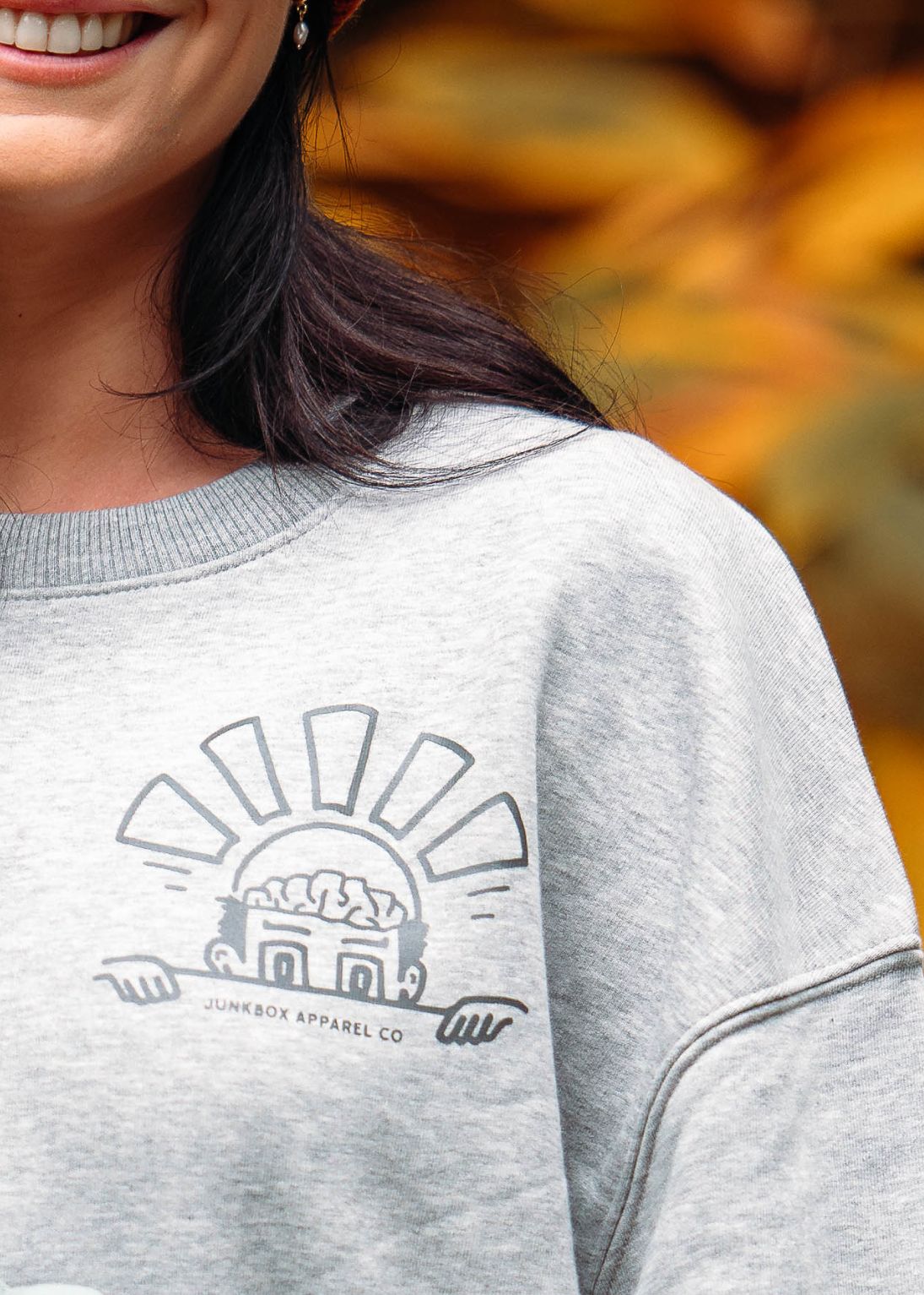 Person wearing a gray sweatshirt with a logo, blurred autumn background