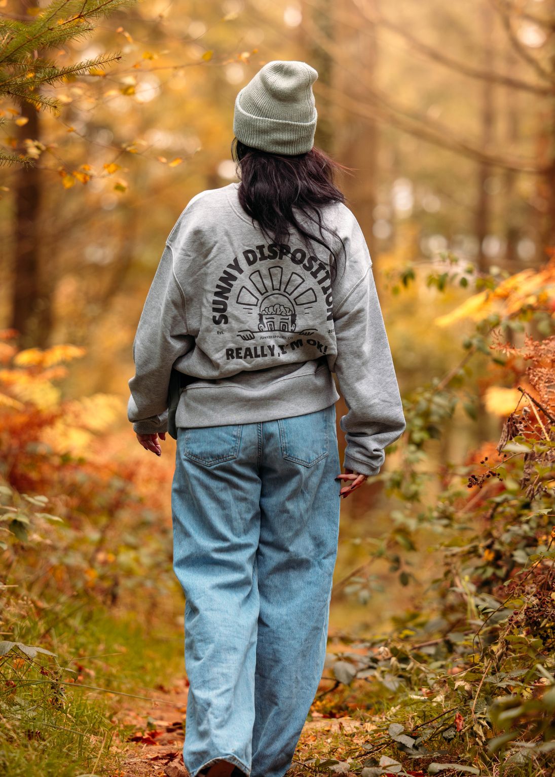 Person wearing a gray sweatshirt with text and a beanie walking through a forest during autumn.